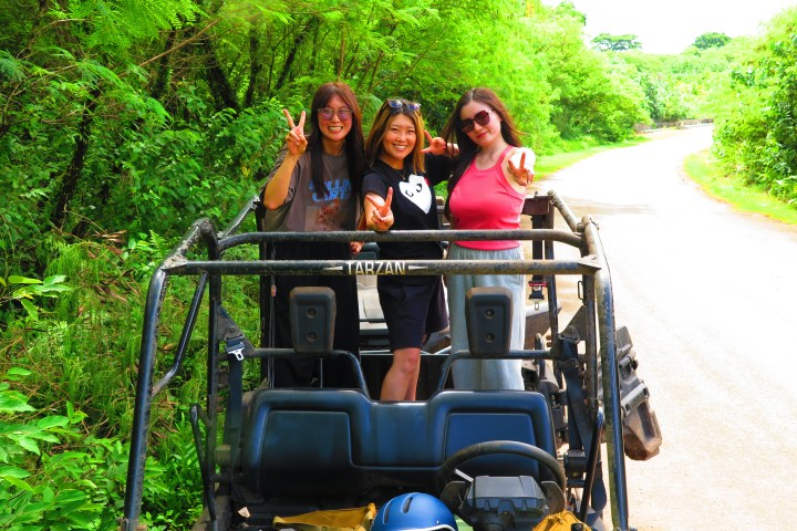 Three people pose in a parked open vehicle on a lush, green roadside.
