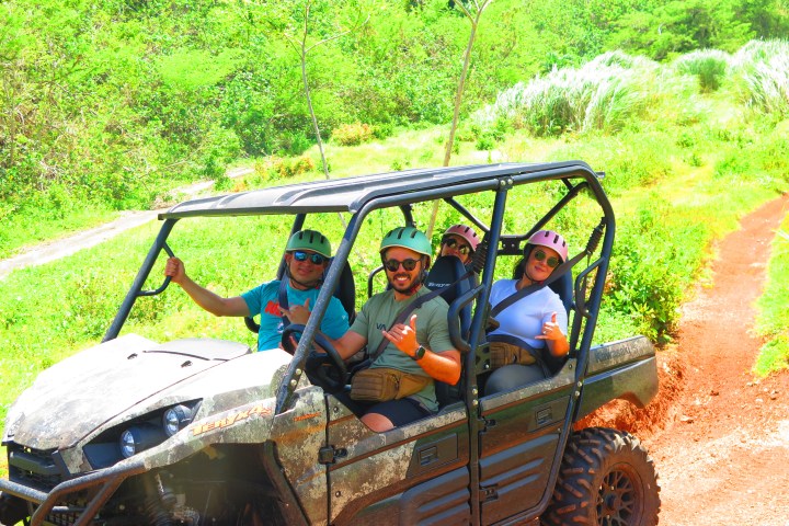 Four people in helmets riding an off-road vehicle on a dirt path surrounded by greenery.
