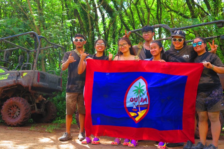 Group holding a Guam flag in a forest beside ATVs.