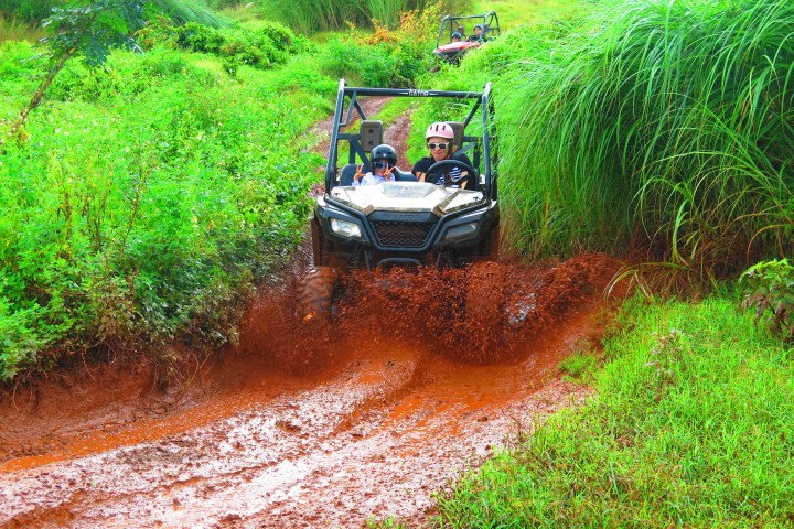 Two people driving an ATV through muddy terrain with greenery.