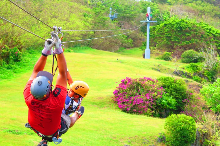 Two people zip-lining over a grassy, hilly landscape with trees and flowers.