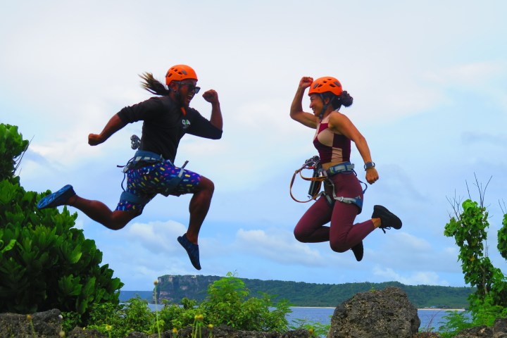 Two people in helmets jumping energetically outdoors with a scenic background.