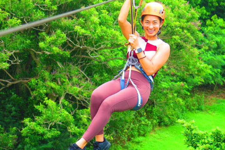 Person zip-lining in harness and helmet over lush green forest.