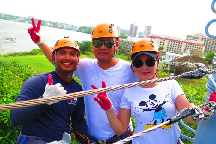 Three people in helmets posing for a photo on a zip line platform overlooking a city and water.