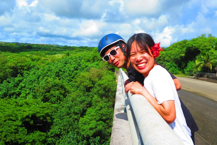 Two people smiling and leaning over a railing with greenery in the background.