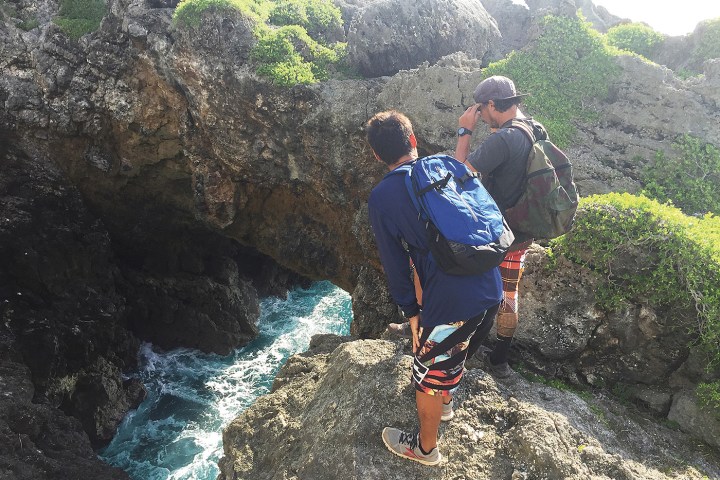Two hikers with backpacks stand on rocky cliff overlooking ocean.