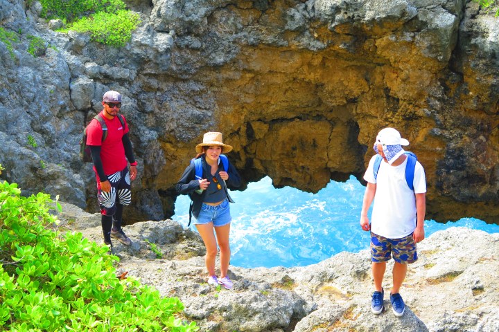 Three hikers stand near a rock formation with a hole, overlooking the ocean below.