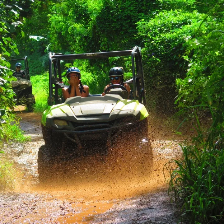 Two people driving an off-road vehicle through a muddy forest path.