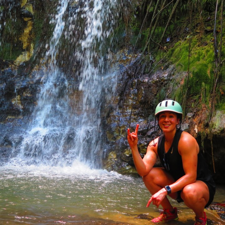 Person in helmet poses by a waterfall, making a hand gesture.