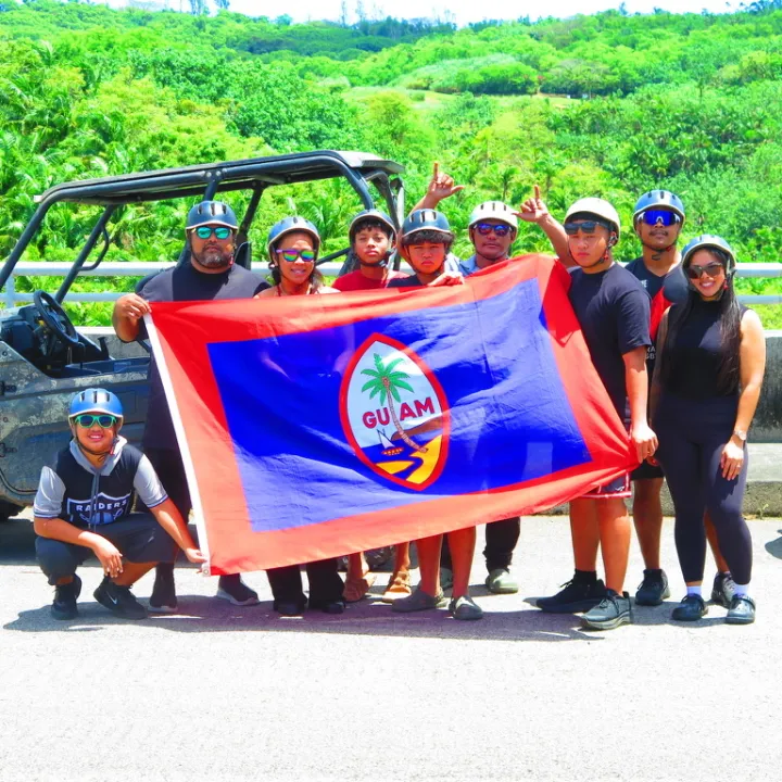 Group holding Guam flag in front of off-road vehicles, with lush greenery in background.