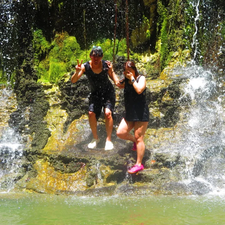 Two people posing by a waterfall with mossy rocks.