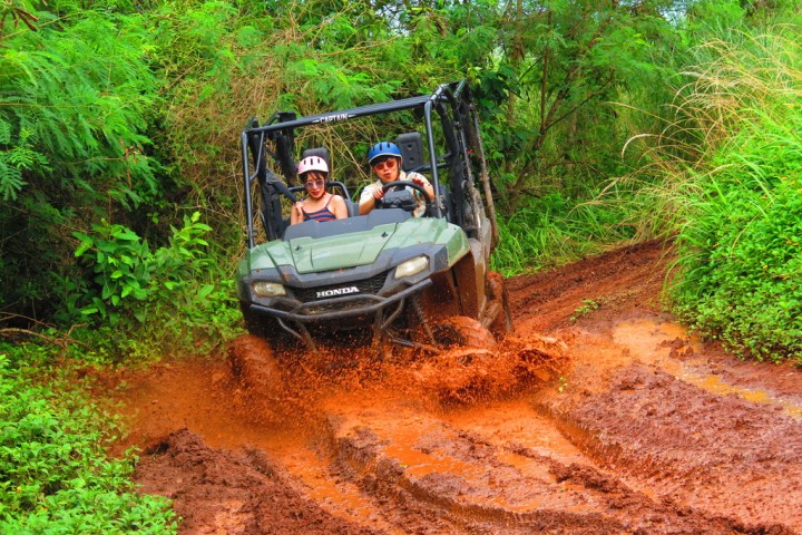 Two people in a green ATV driving through a muddy trail surrounded by lush greenery.