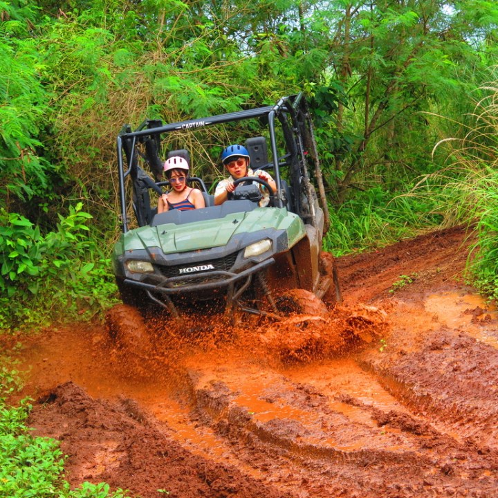 Two people in a green ATV driving through a muddy trail surrounded by lush greenery.