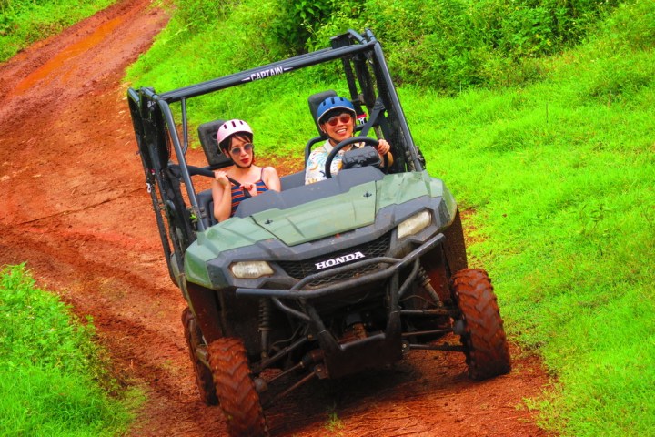 Two people in a green off-road vehicle driving on a muddy path surrounded by grass.