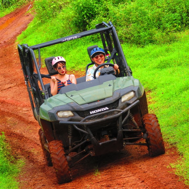 Two people in a green off-road vehicle driving on a muddy path surrounded by grass.