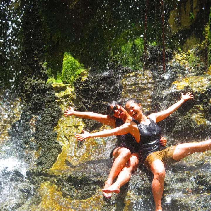 Two people smiling, sitting playfully under a waterfall.