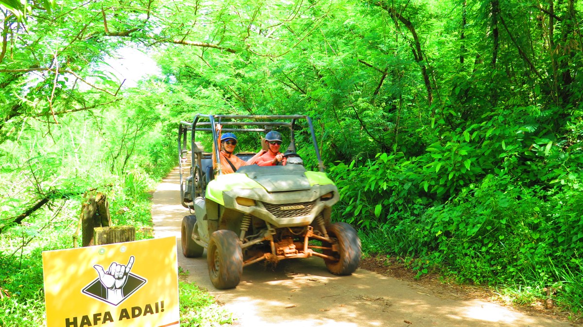 Two people on an ATV on a lush, green forest trail with a 'HAFA ADAI' sign.