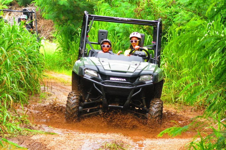 Two people in a green ATV driving through a muddy trail surrounded by lush greenery.