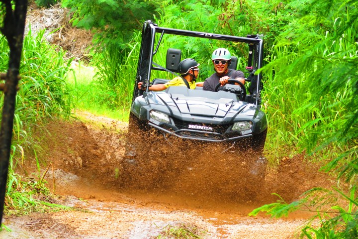 Two people in an ATV driving through a muddy trail surrounded by lush greenery.