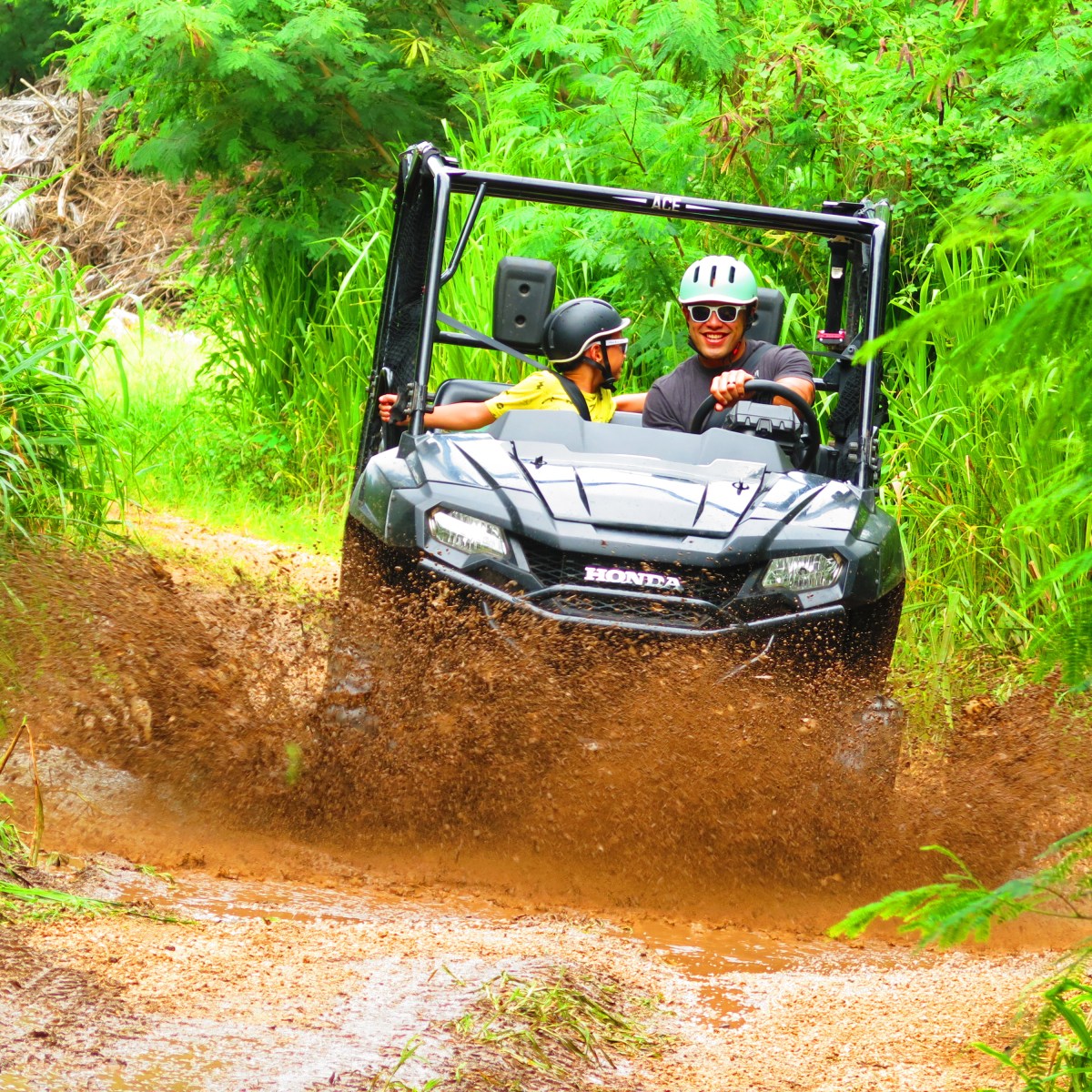 Two people in an ATV driving through a muddy trail surrounded by lush greenery.