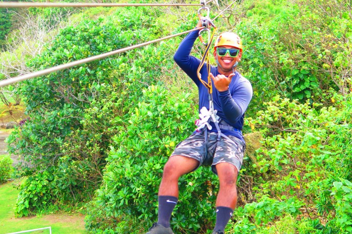 Person ziplining over lush greenery, wearing a helmet and sunglasses, smiling.