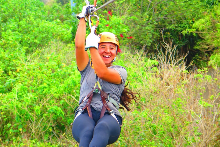Person ziplining with a green forest background, smiling and wearing a helmet.