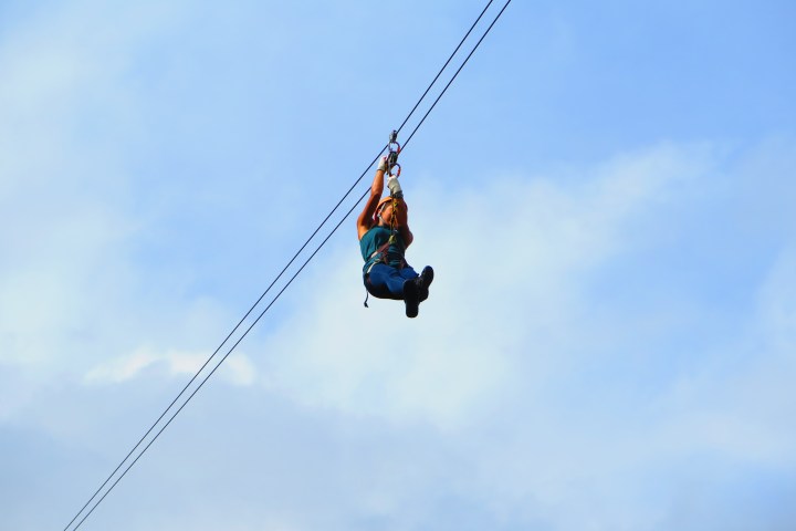 Person zip lining against a clear blue sky with some clouds.