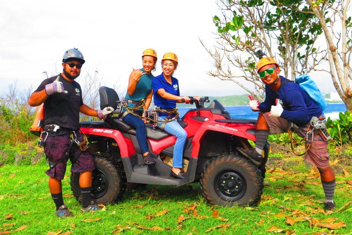 Four people in helmets pose with an ATV on a grassy area by the water.
