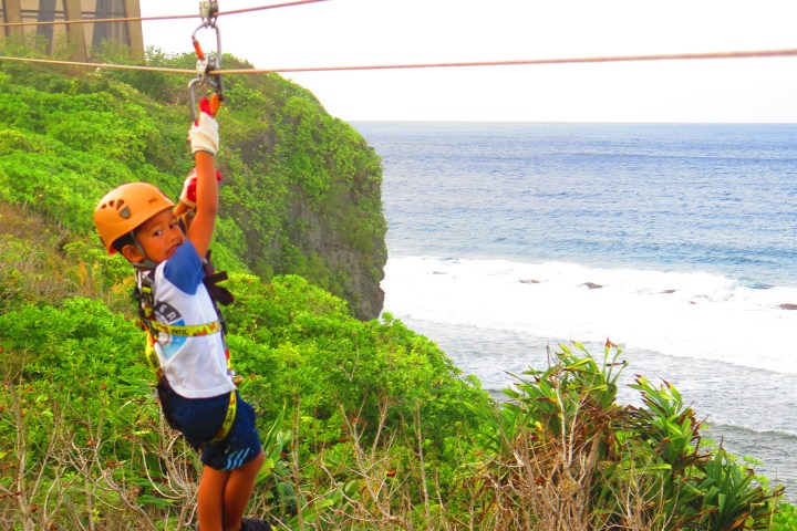 Child on a zip line over a coastal landscape with sea and greenery.