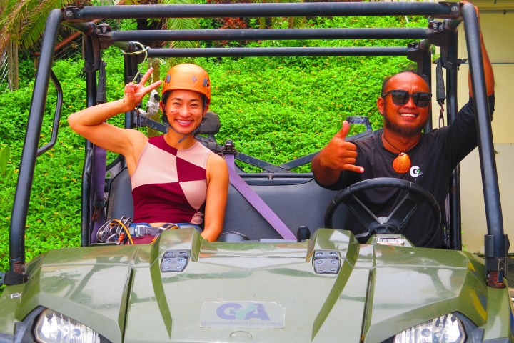 Two people smiling in an off-road vehicle, one giving a peace sign, both wearing helmets.